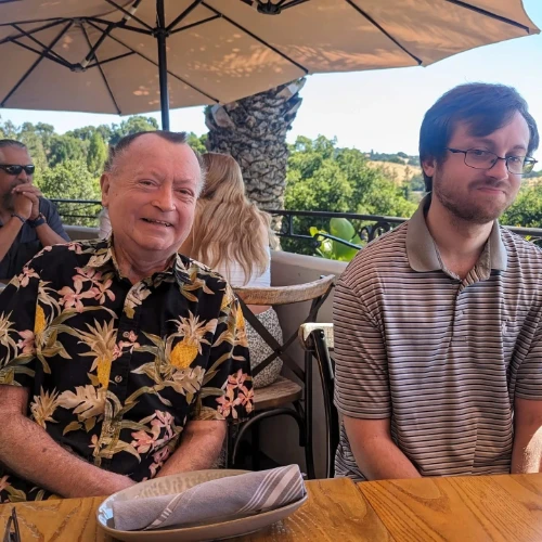 Two people sit at a wooden table outdoors under a large umbrella. On the left, Dave, wearing a floral shirt, smiles warmly. Beside him, the younger individual in a striped shirt maintains a neutral expression. Its the 16th of June, and trees fill the serene background.
