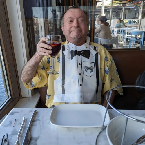 Dave, sitting at a restaurant table next to a window, beams as he holds up a glass of red wine. Sporting a yellow shirt with a tuxedo-themed bib, hes surrounded by empty plates and cutlery at his favorite crab house, which exudes a casual and bright atmosphere.