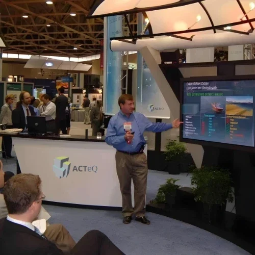 Dave Ridyard is giving a presentation at a trade show booth, with a large monitor displaying graphs and data. The booth showcases ACTeQ branding, and several attendees are watching and engaging in the event.