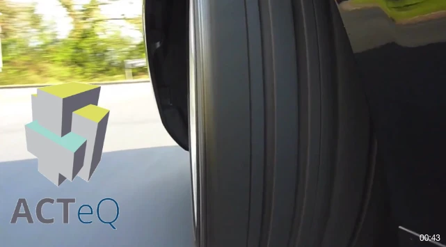 Close-up view of a tire as a vehicle moves on a road, with blurred greenery in the background. A logo with the text ACTeQ and abstract geometric shapes is visible on the left side of the image.