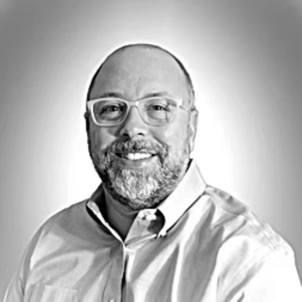 Black and white portrait of Brandon Mattox, a bearded man wearing glasses and a light-colored button-up shirt, smiling against a gradient background.